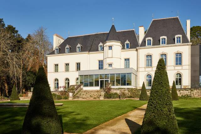 Château du Domaine du Liziec avec façade claire, tourelle centrale, jardin paysager et allée en pierre sous ciel bleu