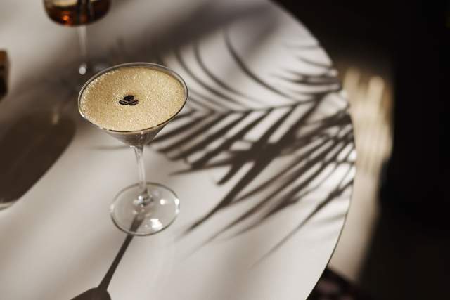 Cocktail raffiné sur surface blanche au Domaine du Liziec, ombre de feuilles tropicales créant une ambiance estivale et élégante