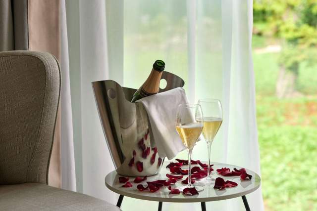 Table romantique au Domaine du Liziec avec champagne, pétales de rose et vue sur la verdure à travers des rideaux blancs