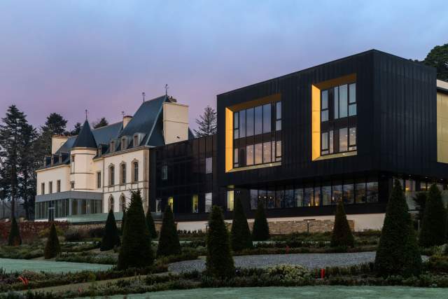 Vue du Domaine du Liziec avec bâtiment moderne aux accents jaunes et manoir traditionnel, entourés de jardins paysagers au lever ou coucher du soleil