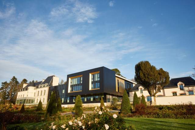 Architecture du Domaine du Liziec avec bâtiment moderne noir à grandes baies vitrées et bâtisse traditionnelle, entourés de jardins fleuris sous ciel clair