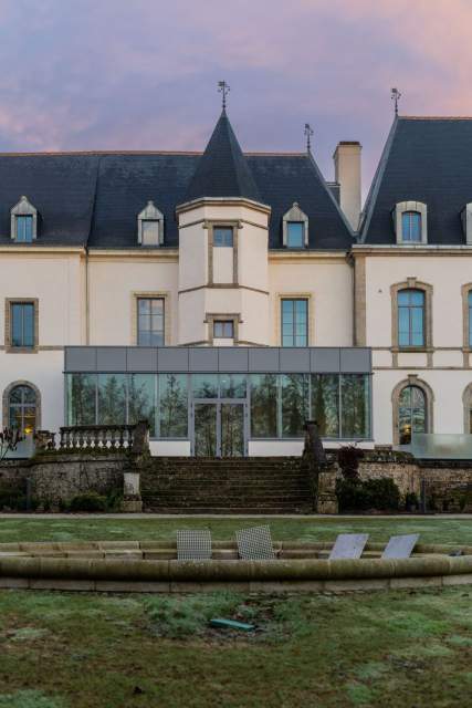 Vue du Domaine du Liziec avec bâtiment élégant à tourelle centrale, verrière, jardin aménagé et fontaine entourée de transats